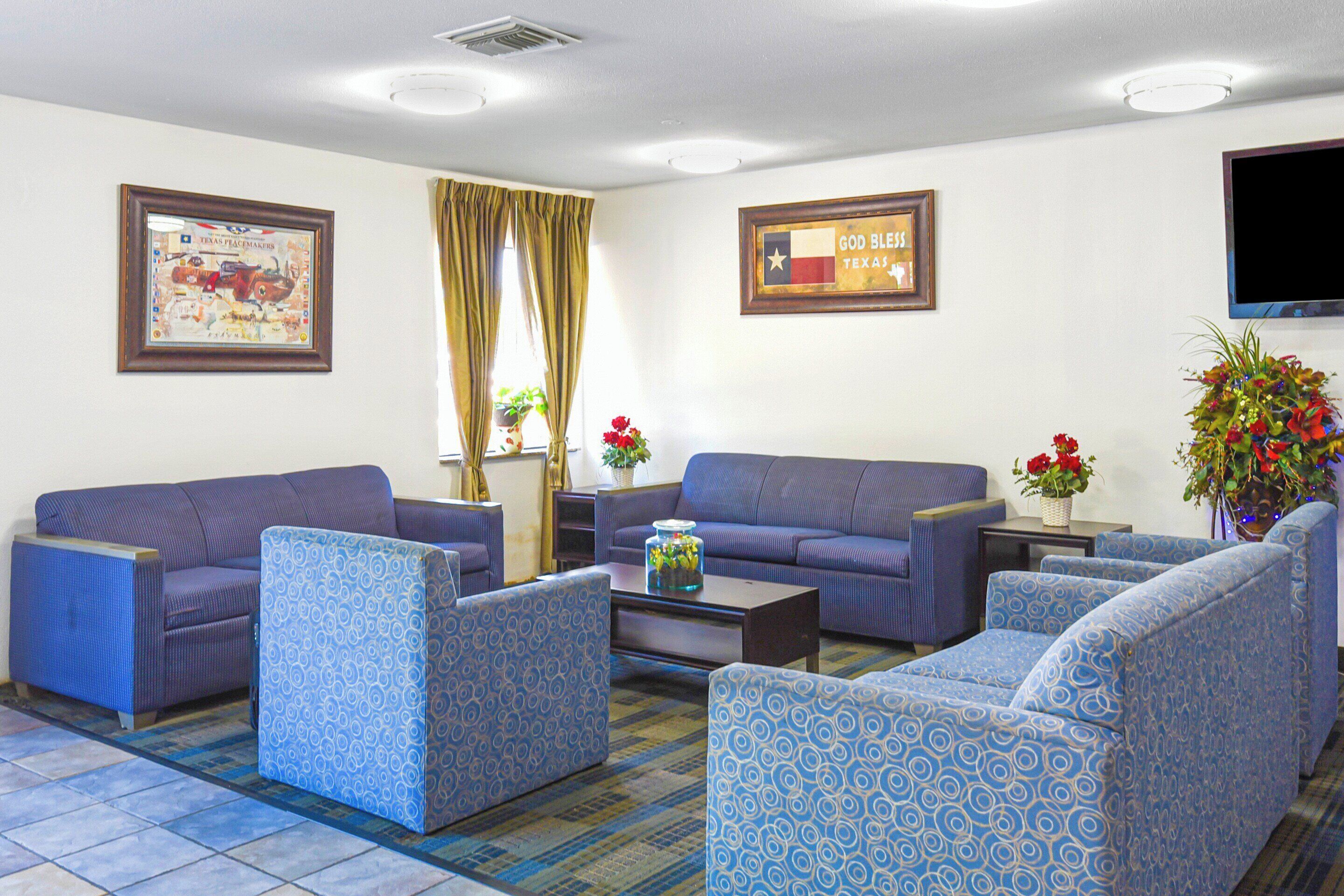 A hotel room with a blue patterned sofa set, a flat-screen TV mounted on the wall, and a framed Texas flag.