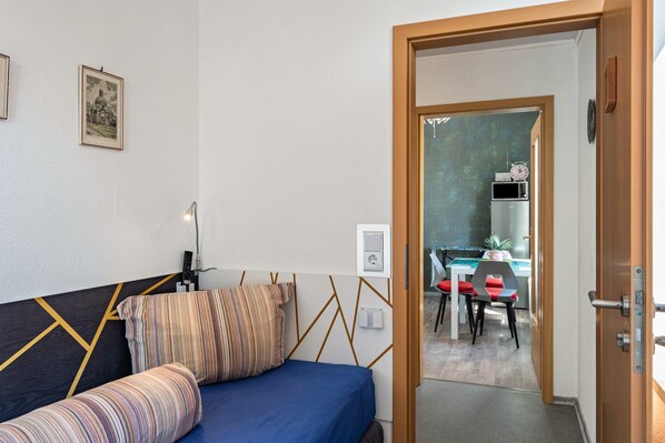 A bedroom with a bed, a bedside table, a lamp, and a door leading to a dining area.