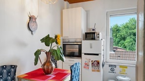 Una cucina compatta con forno a microonde, forno e frigorifero, una sveglia al muro e una finestra vista sul verde.