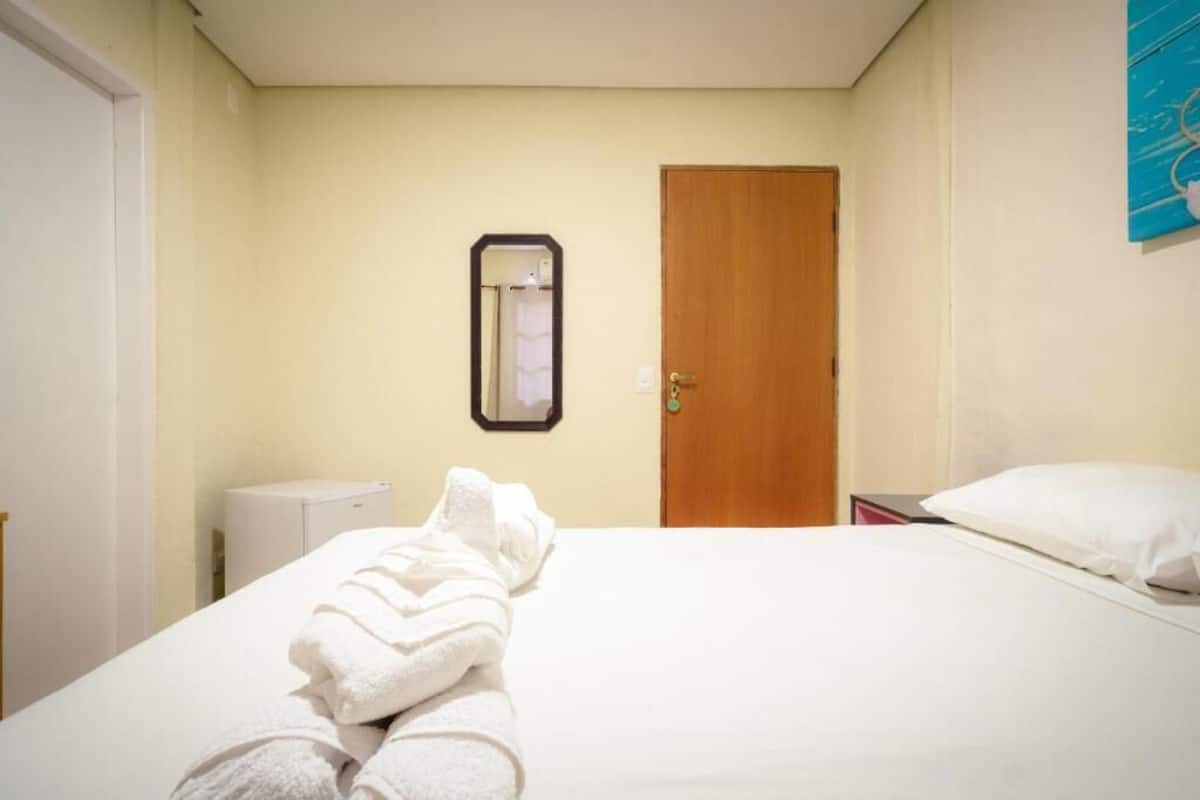 A hotel room with a bed, a wooden door, a small white cabinet, and a mirror on the wall.
