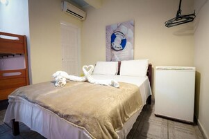 Hotelkamer met een bed, een witte muur, een deur, een wandairconditioner, een kleine witte koelkast en beddecoratie.