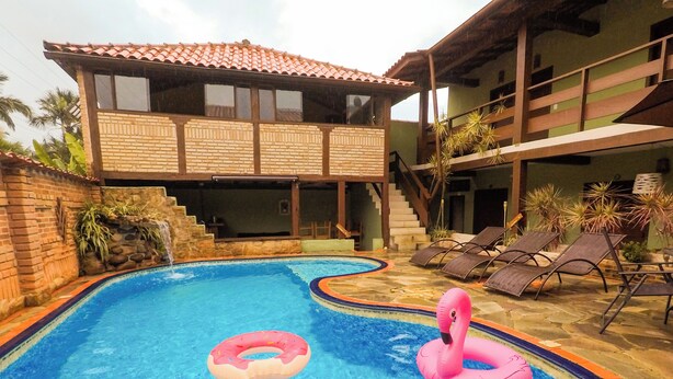 A swimming pool with two pink inflatable rings, a brick wall, and a two-story building with a tiled roof. - VELINN Pousada Kyrios (São Sebastião)