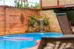 Una piscina con un muro de ladrillo, un muro de piedra con cascada, y una silla de descanso.