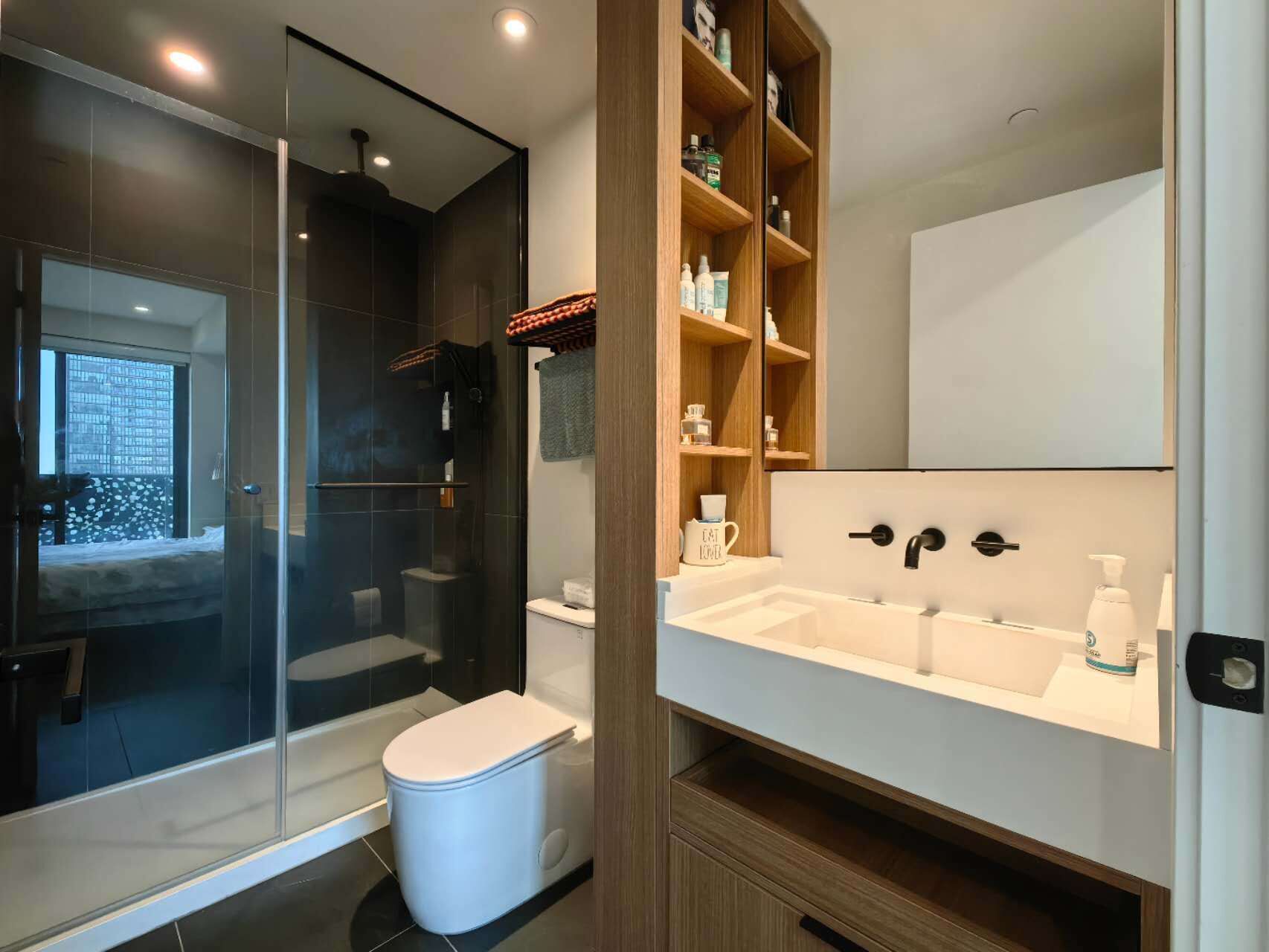 Une salle de bain moderne dotée d’une douche à parois de verre, de toilettes, d’un lavabo avec un miroir et d’une étagère en bois où sont rangés les articles de toilette.