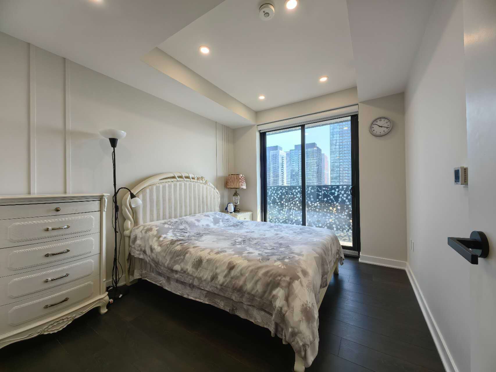 Un dormitorio con cama, tocador, lámpara y vistas a edificios a través de un ventanal grande.