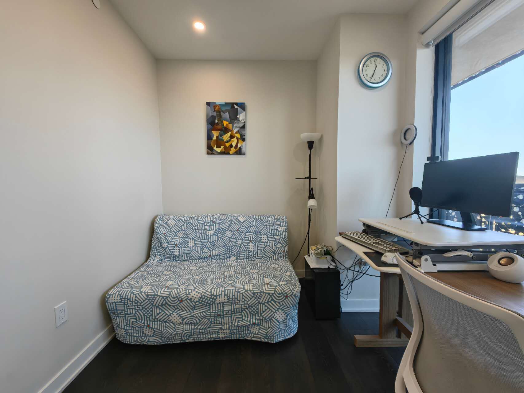 Ein kleiner Raum mit einem Bett, einem Schreibtisch mit Computer, einer Uhr und einem Fenster.