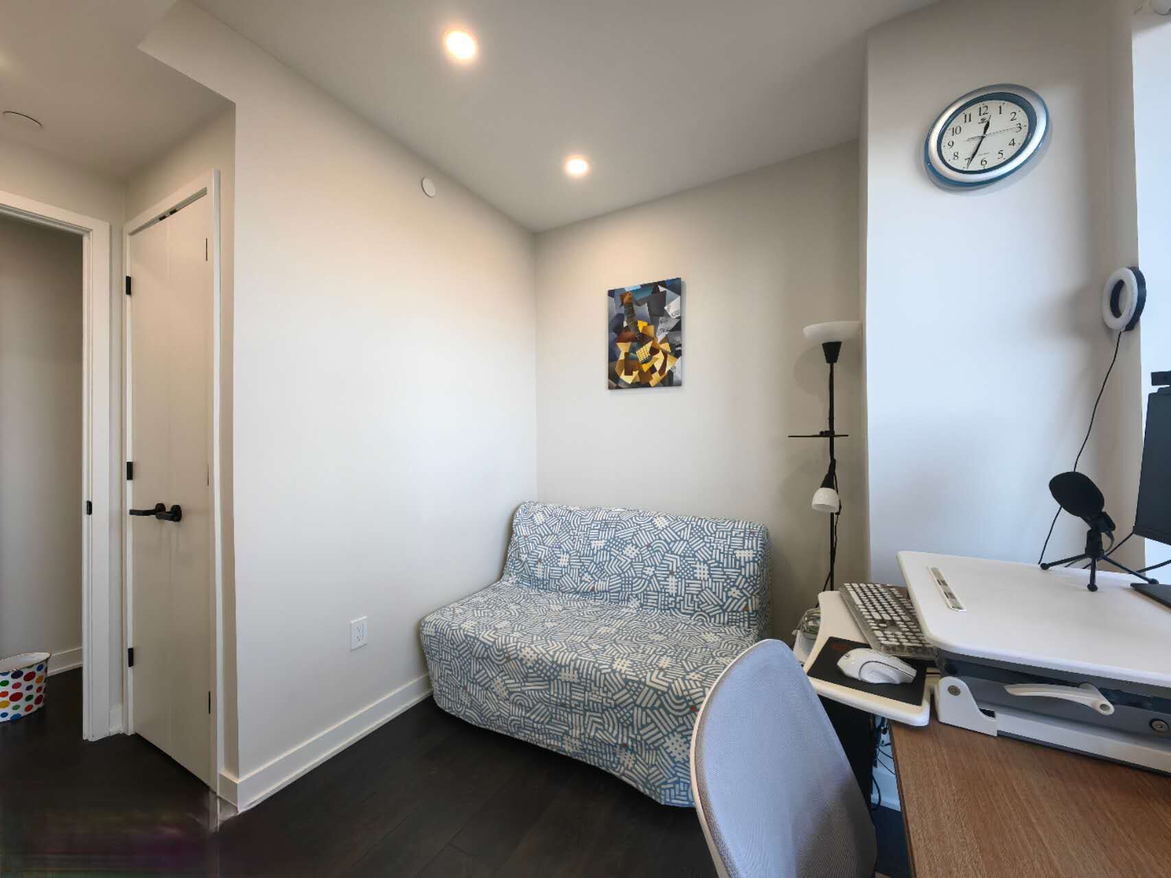 A small, modern room with a patterned sofa, a desk with a computer, a clock, and a painting on the wall.