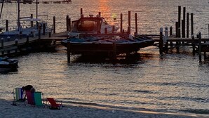 Un coucher de soleil sur une marina où sont amarrés des bateaux, une jetée qui s’avance dans l’eau et des chaises de plage sur le sable.