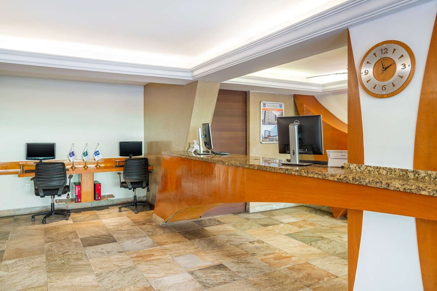 Un área de recepción con mostrador de madera, dos monitores de computadora y un reloj en la pared.