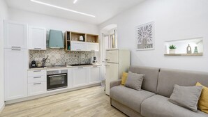 Una cucina moderna con mobili bianchi, forno integrato e un divano grigio con cuscini.