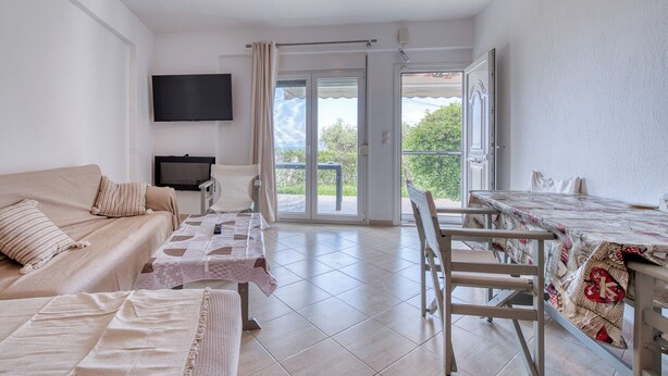 Une pièce comprenant un canapé, une table recouverte d’une nappe, un téléviseur et une porte-fenêtre coulissante donnant sur un espace extérieur.