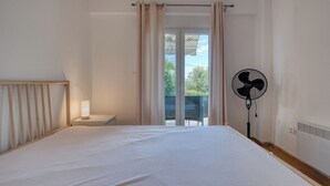 Une chambre à coucher comprenant un lit, une table de chevet, un ventilateur sur pied et une vue sur l’extérieur grâce à une porte coulissante.