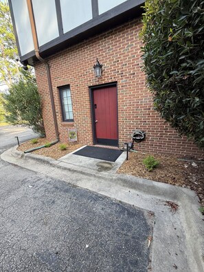 A brick building with a red door, a small window, and a black awning over the entrance. - Experience the charm of Manteo from this inviting 2 bedroom apartment. (Manteo)