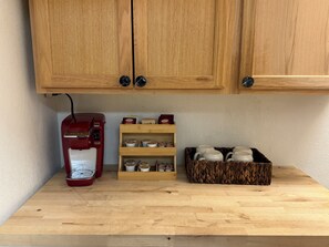 A wooden kitchen counter with a red coffee maker, a shelf with tea and coffee boxes, and a basket with white mugs. - Experience the charm of Manteo from this inviting 2 bedroom apartment. (Manteo)