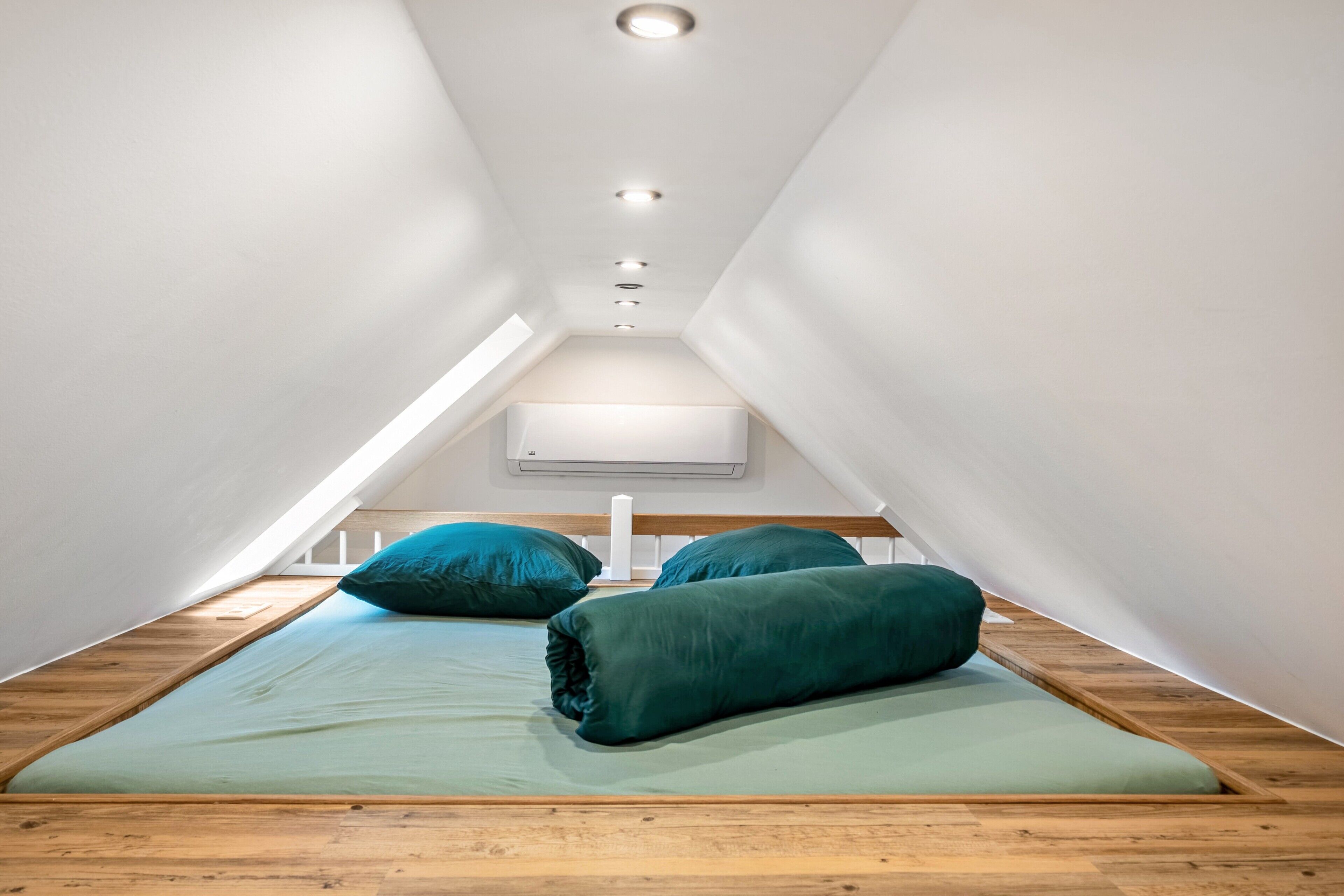 Une chambre avec un plafond en pente, un lit avec une literie verte et un climatiseur blanc fixé au mur.