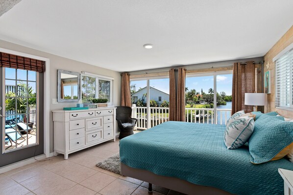 A bedroom with a large bed, a dresser, a chair, and a balcony with a view.