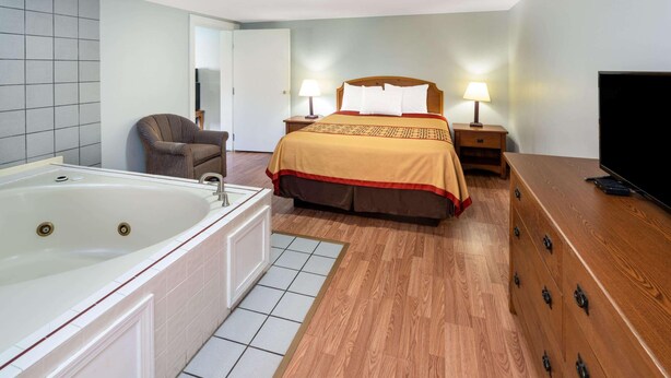 Una habitación de hotel con una bañera grande, una cama con cubrecama rojo y marrón, dos mesitas de noche con lámparas, un aparador de madera y un televisor.