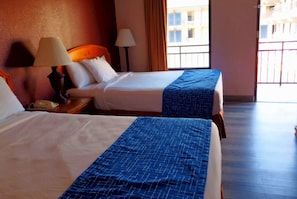 Two beds with white and blue bedding, a wooden headboard, a nightstand with a lamp, and a view of buildings through a window. - Travelodge by Wyndham Pigeon Forge (Pigeon Forge)