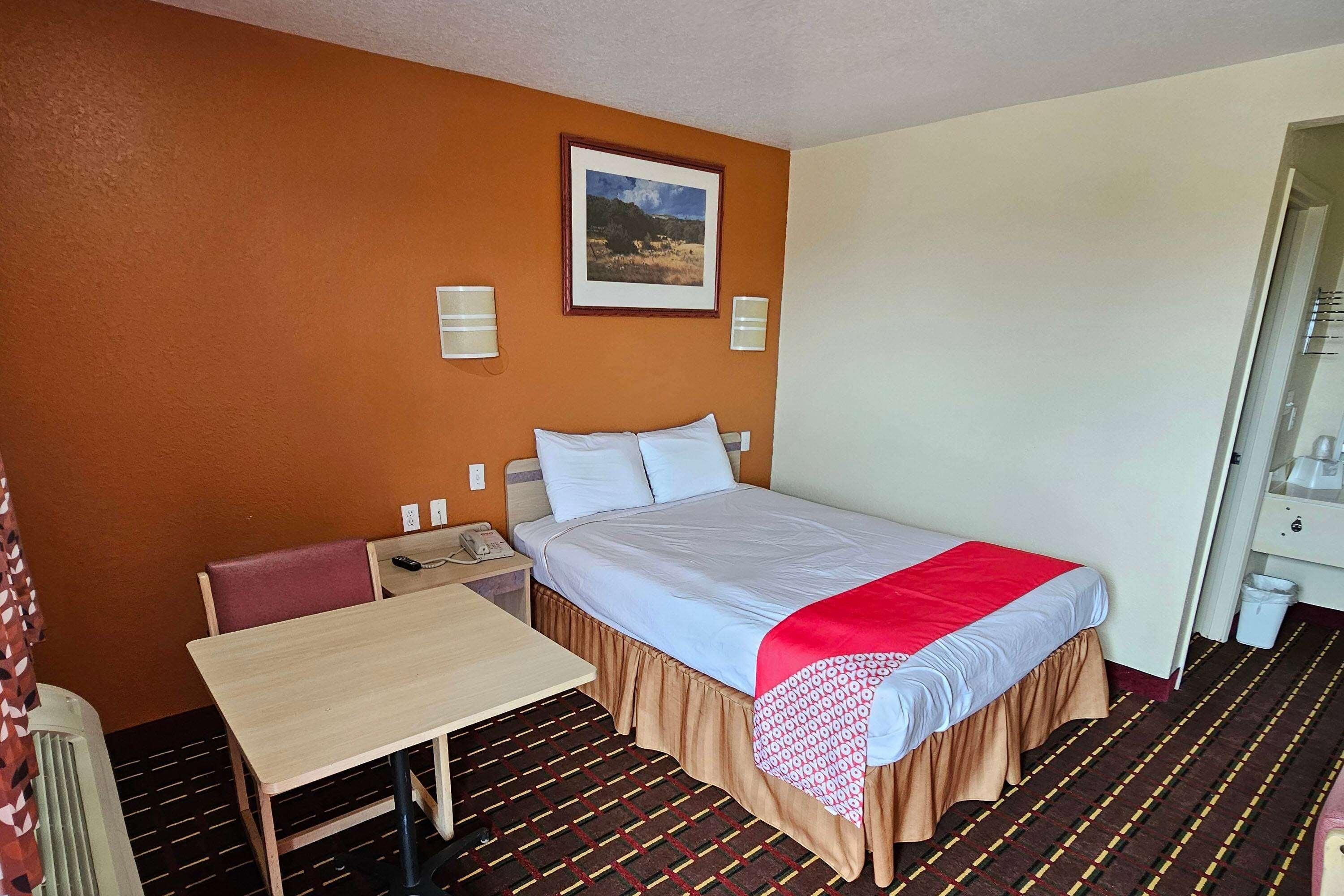 A hotel room with a bed, a table, a chair, a nightstand, a telephone, and a framed picture on the wall.