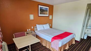 A hotel room with a bed, a table, a chair, a nightstand, a telephone, and a framed picture on the wall.