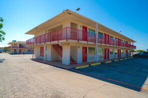 Un motel de dos pisos con fachada roja y blanca, una zona de estacionamiento cubierta y una escalera visible que conduce al piso superior.