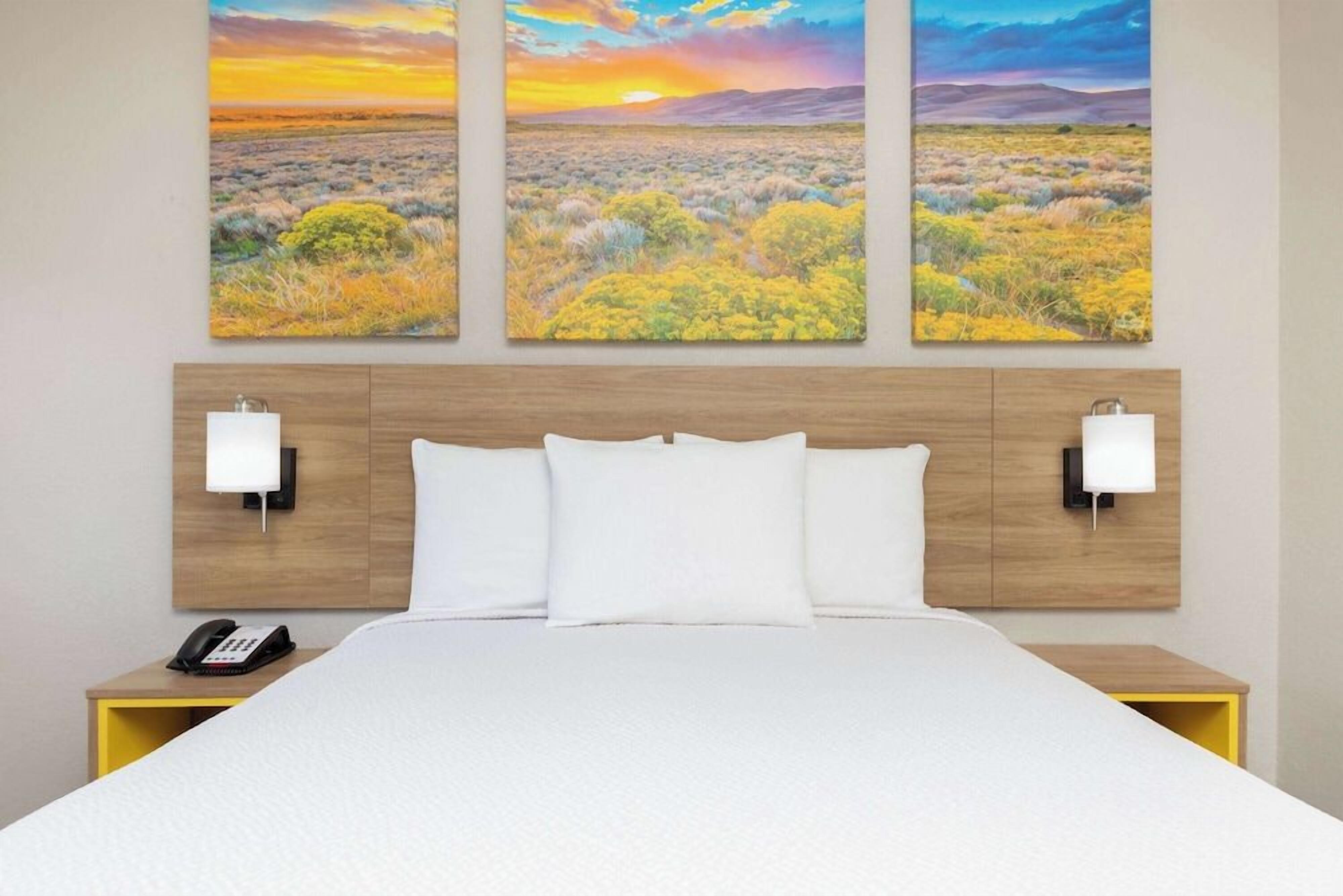 A hotel room with a bed, two bedside lamps, a telephone, and three landscape paintings on the wall.