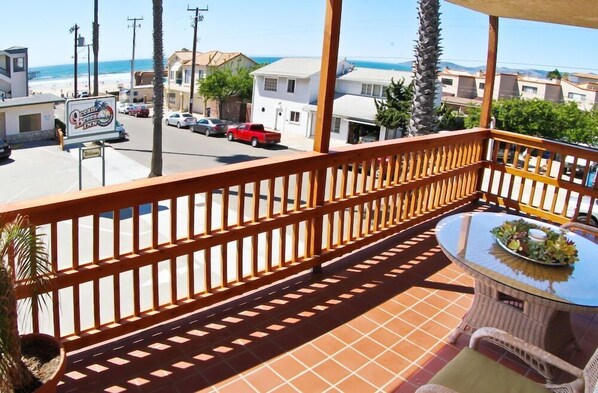 Uma varanda com guarda-corpo de madeira, uma mesa de vidro com planta e vista para a praia e edifícios.