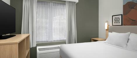 A hotel room with a bed, a TV, a heater, a window with curtains, a nightstand, and a wall art piece.