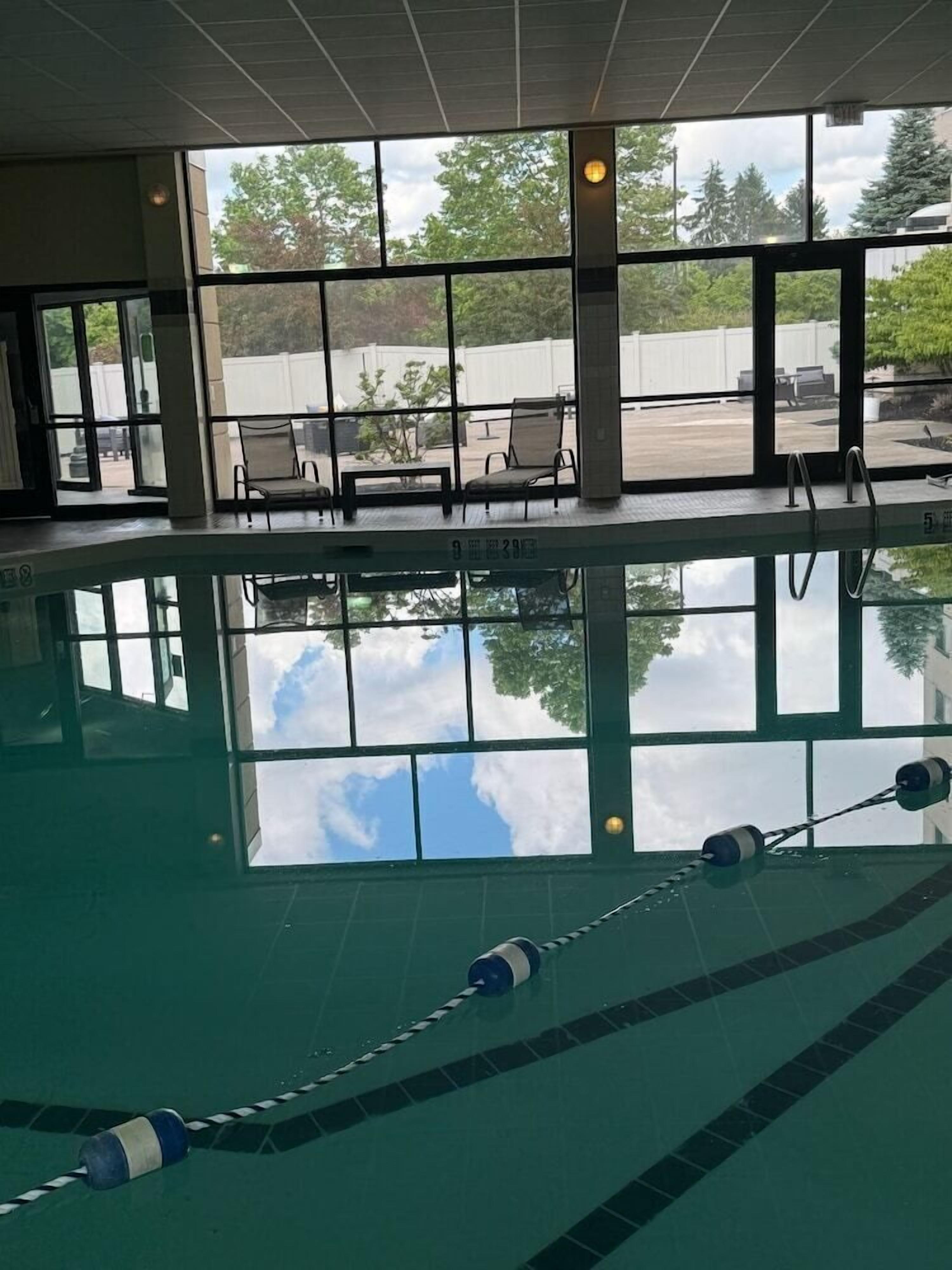 An indoor swimming pool with clear glass walls, a view of the sky, and a seating area with chairs.