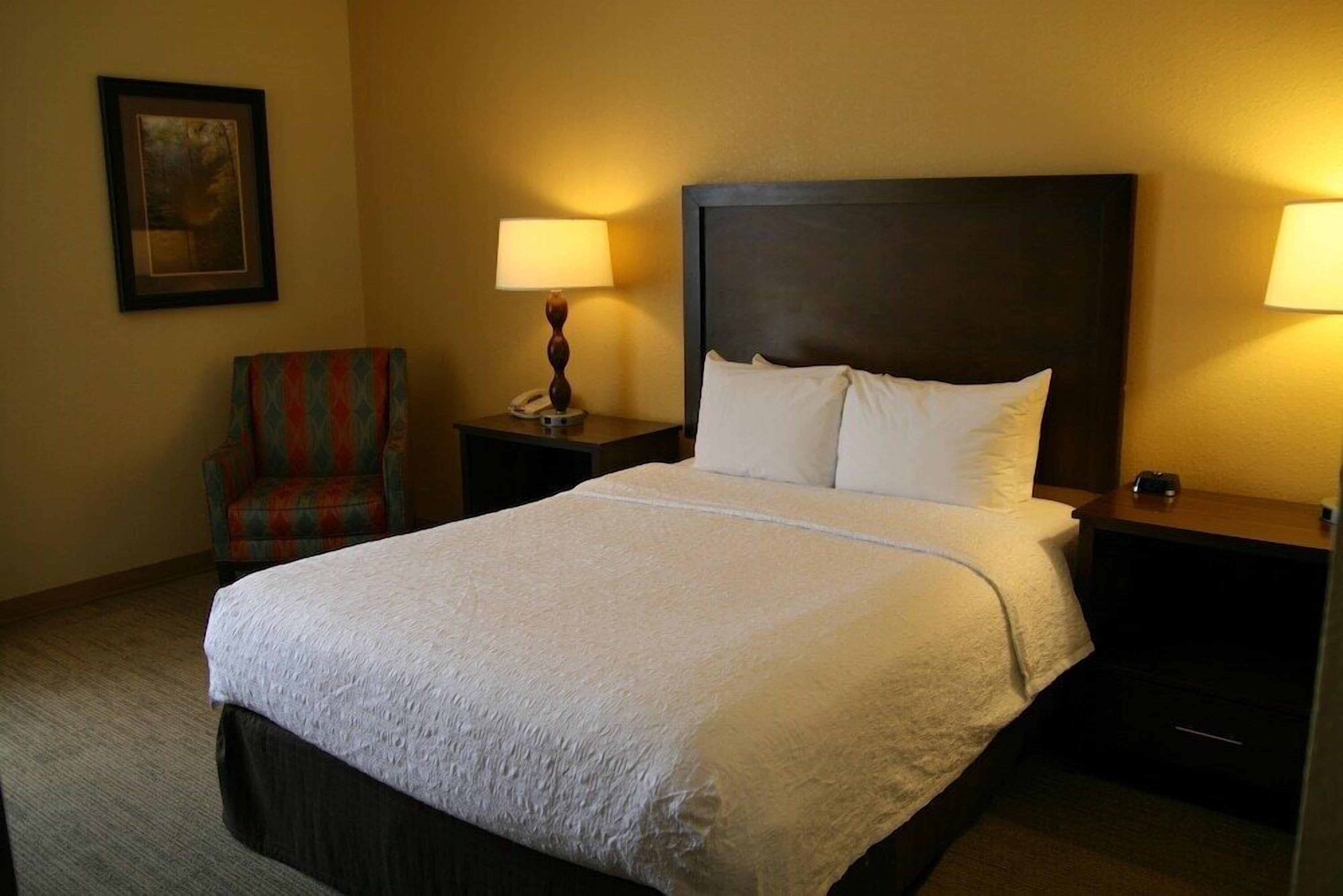 A hotel room with a large bed, two bedside tables, a chair, and a framed picture on the wall.