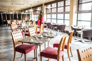 A restaurant with multiple tables and chairs, large windows, and potted plants.