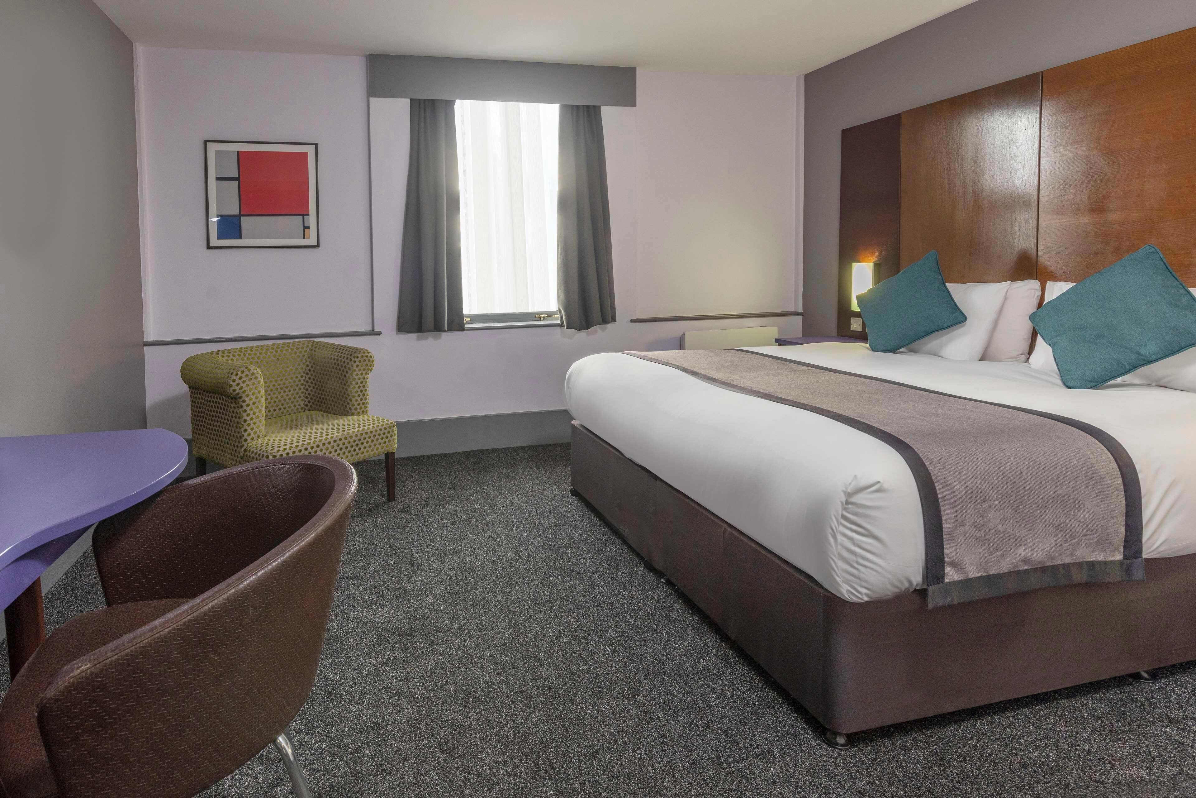 A hotel room with a bed, a chair, a table, and a window with curtains.