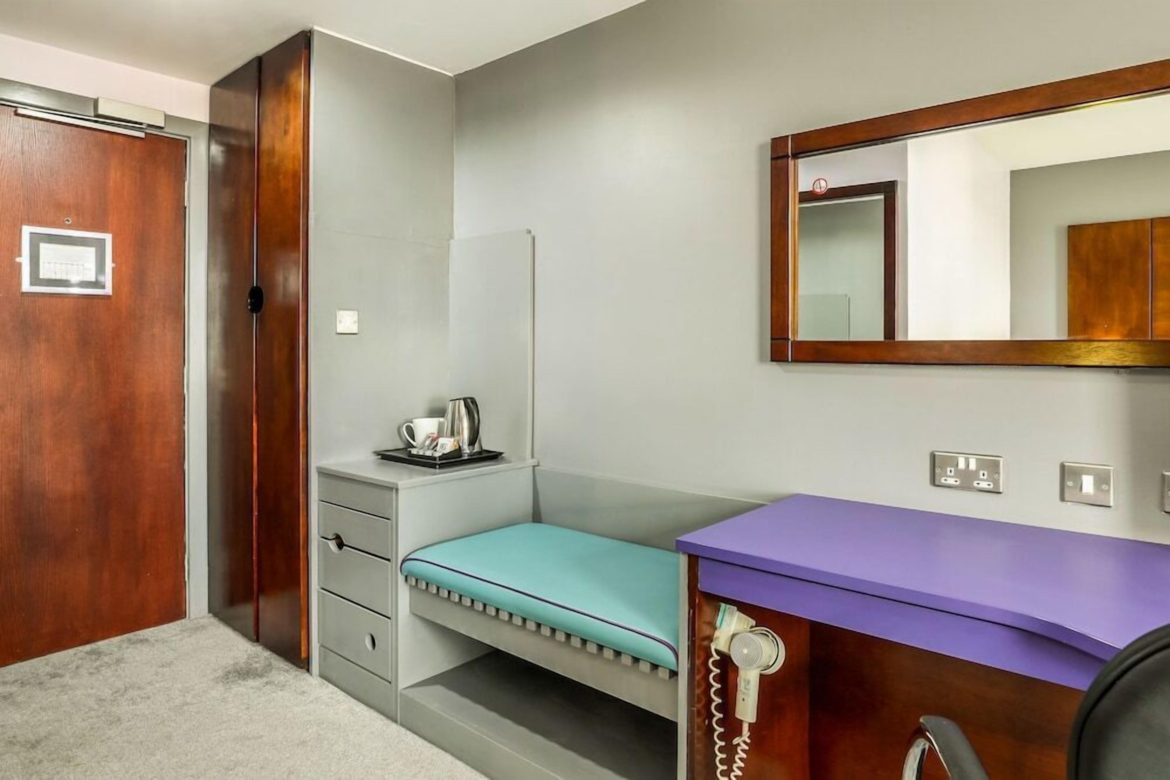 Quarto com porta de madeira, uma pequena escrivaninha com tampo roxo, um telefone, um espelho e um interruptor de luz.