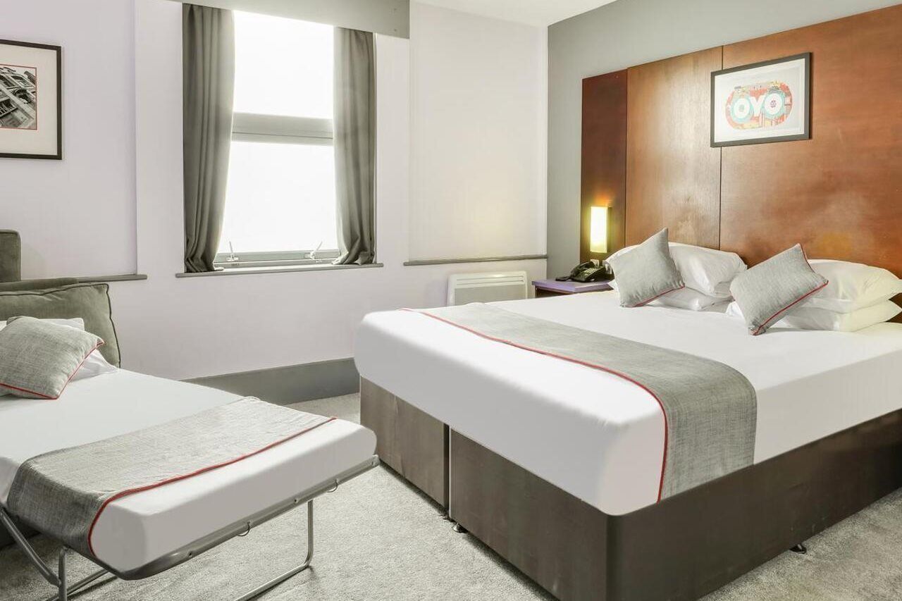 Ett hotellrum med två sängar, ett stort fönster med gardiner, en sänggavel i trä och konstverk på väggarna.