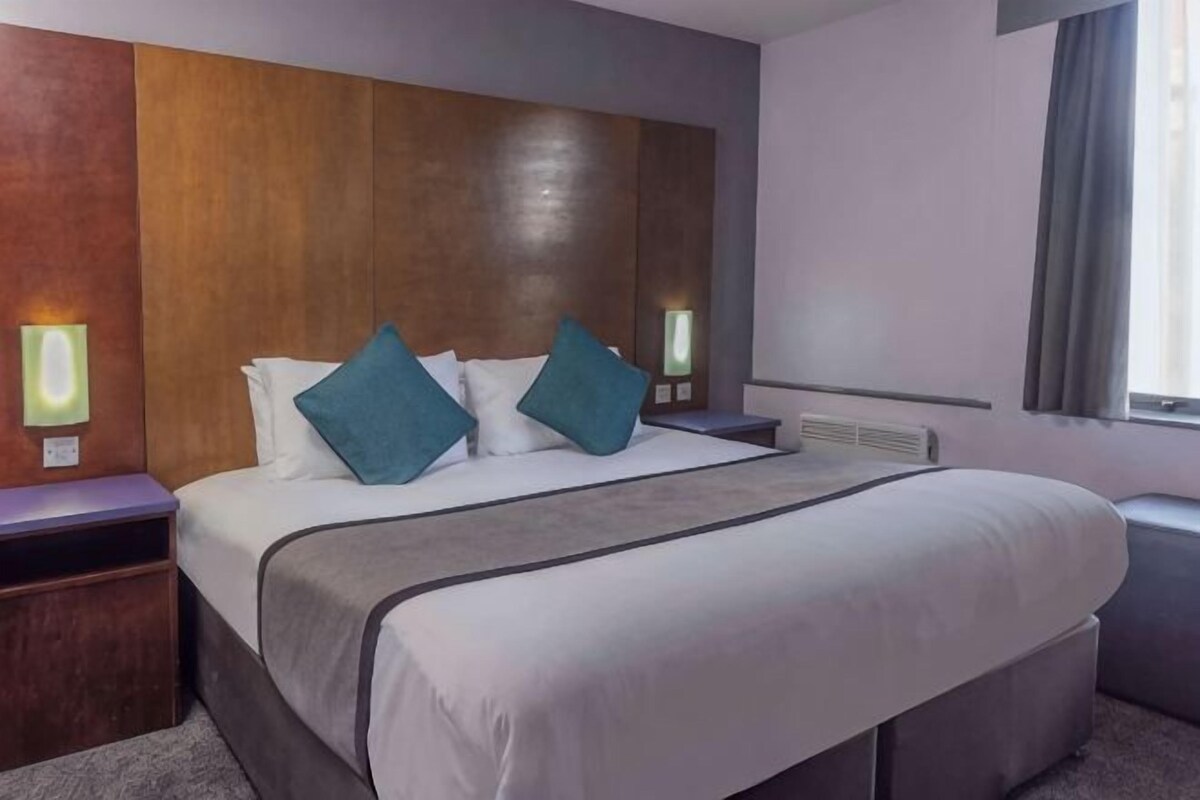 A hotel room with a large bed, two bedside tables, a nightstand, a window with curtains, and wall-mounted lights.
