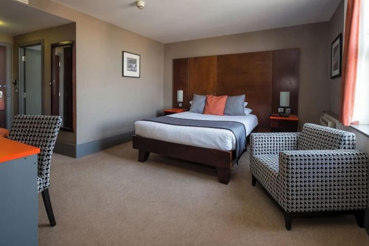 A hotel room with a large bed, two armchairs, a desk, and a door leading to another room.