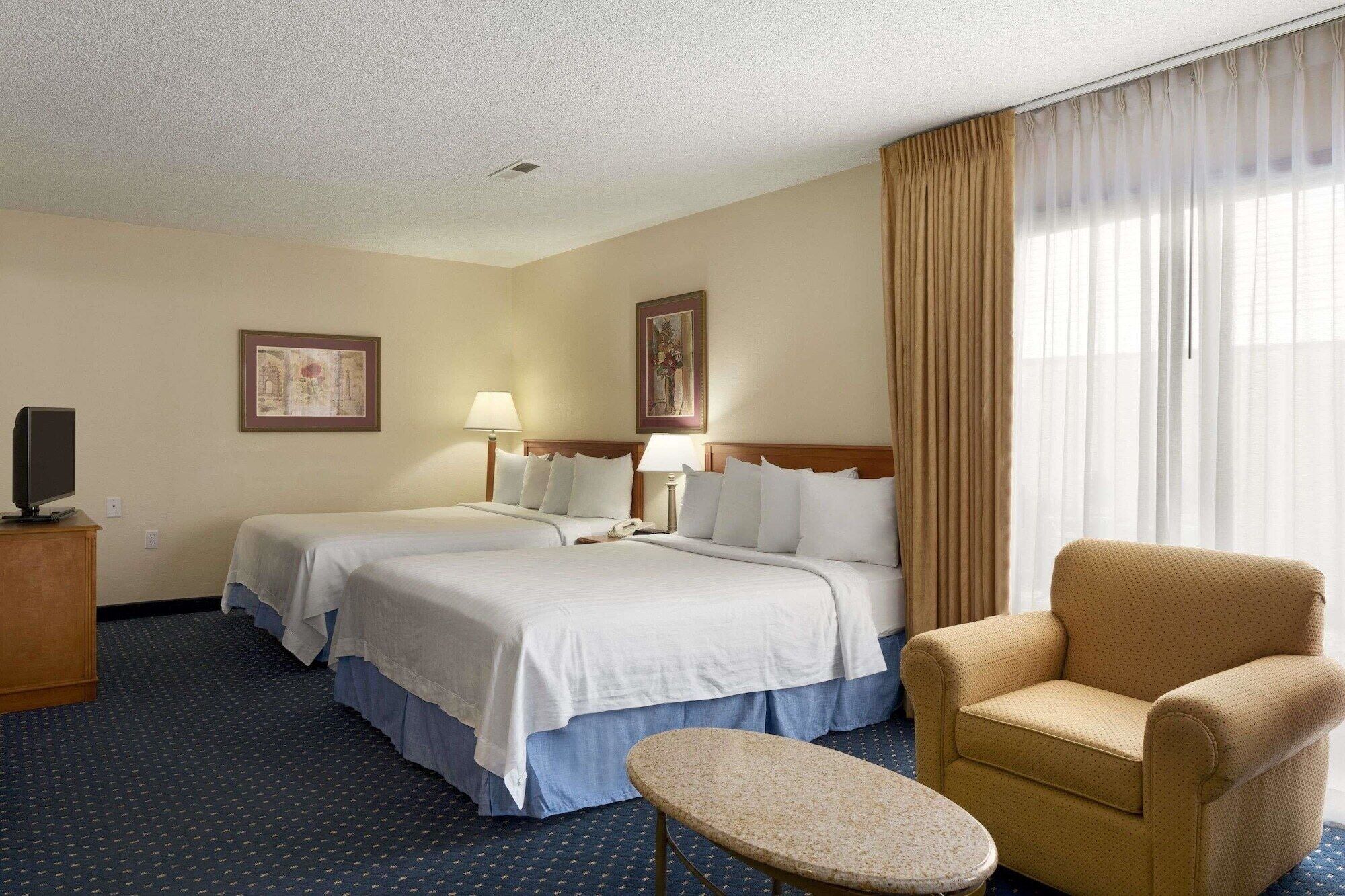 A hotel room with two beds, a chair, a small table, a TV, and artwork on the walls.