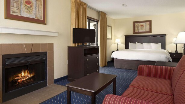 A hotel room with a fireplace, a bed, a desk, a TV, and a large window. - Suburban Studios Fort Wayne, IN (Fort Wayne)