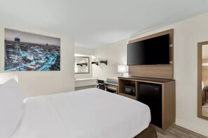 A hotel room with a bed, a flat-screen TV, a desk with a lamp, a microwave, and a cityscape painting. - Clarion Pointe Kennesaw Town Center (Kennesaw)