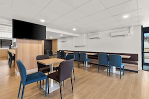 A modern dining area with tables and chairs, a flat-screen TV mounted on a wall, and air conditioning units. - Clarion Pointe Kennesaw Town Center (Kennesaw)
