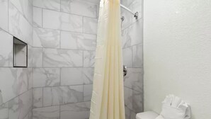 A bathroom with white marble tiles, a shower curtain, and a toilet with a towel rack. - Clarion Pointe Kennesaw Town Center (Kennesaw)
