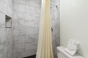 A bathroom with white marble tiles, a shower curtain, and a toilet with a towel rack. - Clarion Pointe Kennesaw Town Center (Kennesaw)