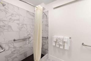 A bathroom with marble tiles, a shower curtain, a towel rack with towels, and a handrail. - Clarion Pointe Kennesaw Town Center (Kennesaw)
