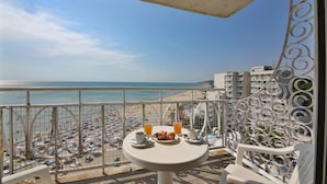 Un balcón con una mesa para dos, con vistas a una playa con numerosas sombrillas y un cielo despejado.