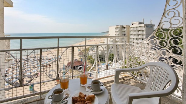 Un balcón con una mesa para dos, con vista a una playa con sombrillas y un edificio que dice 'ELITSA' al fondo.