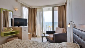 Habitación de hotel con cama, televisión, zona de estar, balcón con vistas al mar y una mesita con un florero.