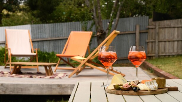 Een terras met twee oranje stoelen, een tafel met een kaasplankje en twee glazen roze drank.