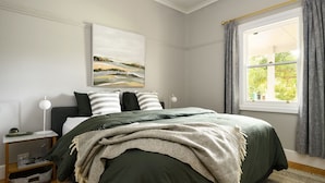 Een slaapkamer met een bed, nachtkastje, lamp, raam met gordijnen en een schilderij aan de muur.