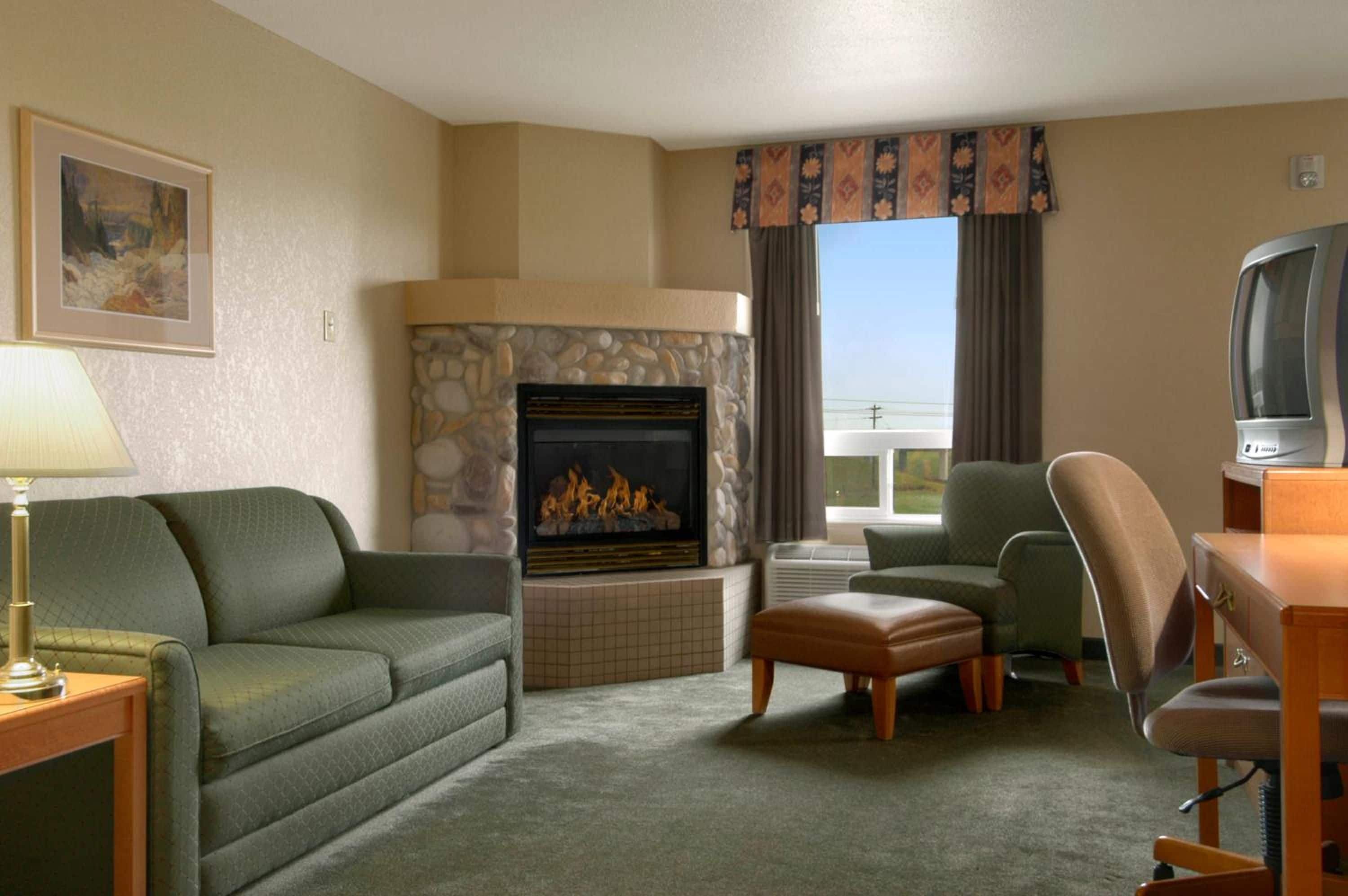 A hotel room with a stone fireplace, green sofa, armchair, ottoman, and a television on a wooden table.