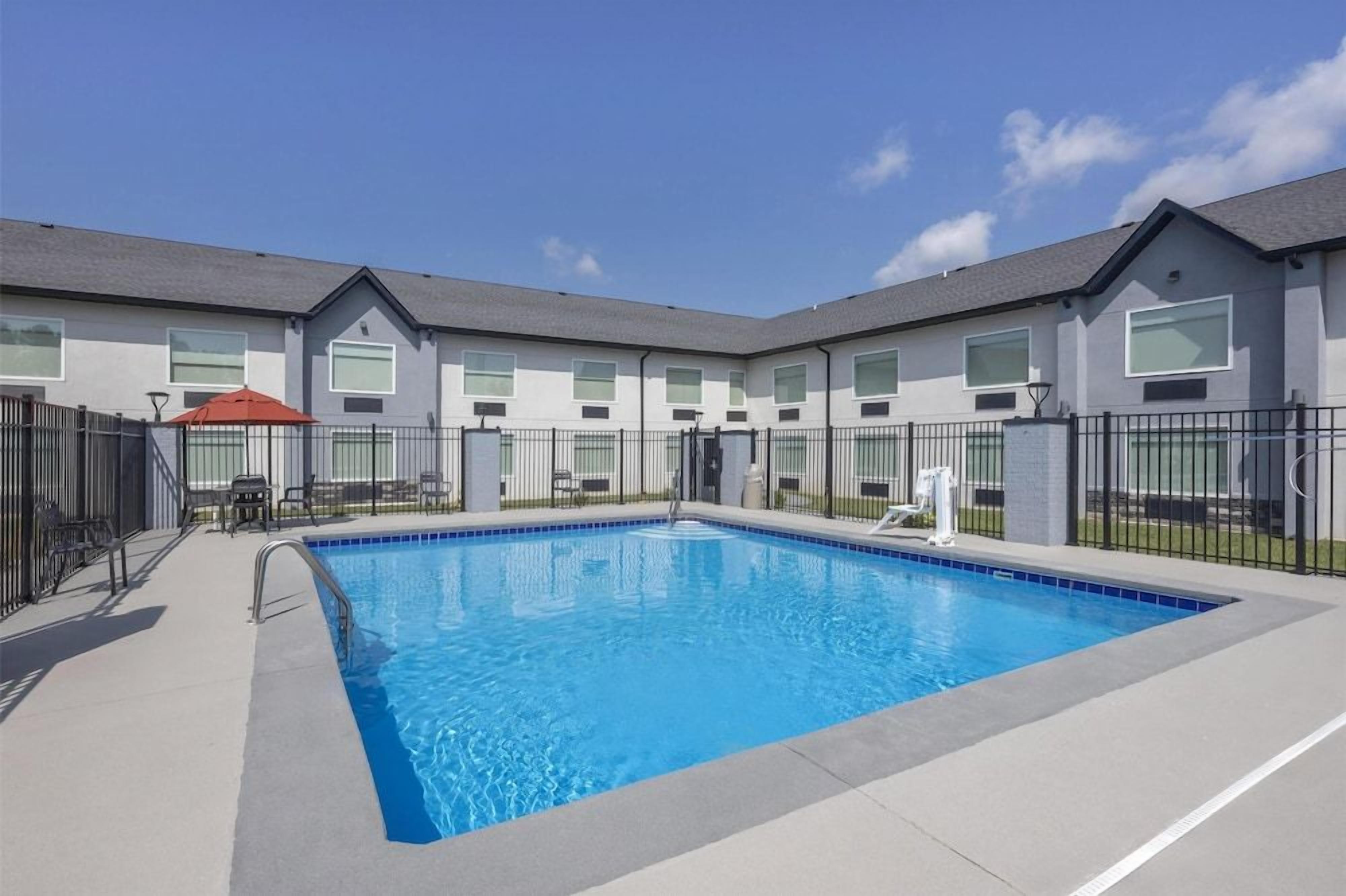 A swimming pool with a clear blue water surface, surrounded by a fence, and adjacent to a building with multiple windows and a flat roof.